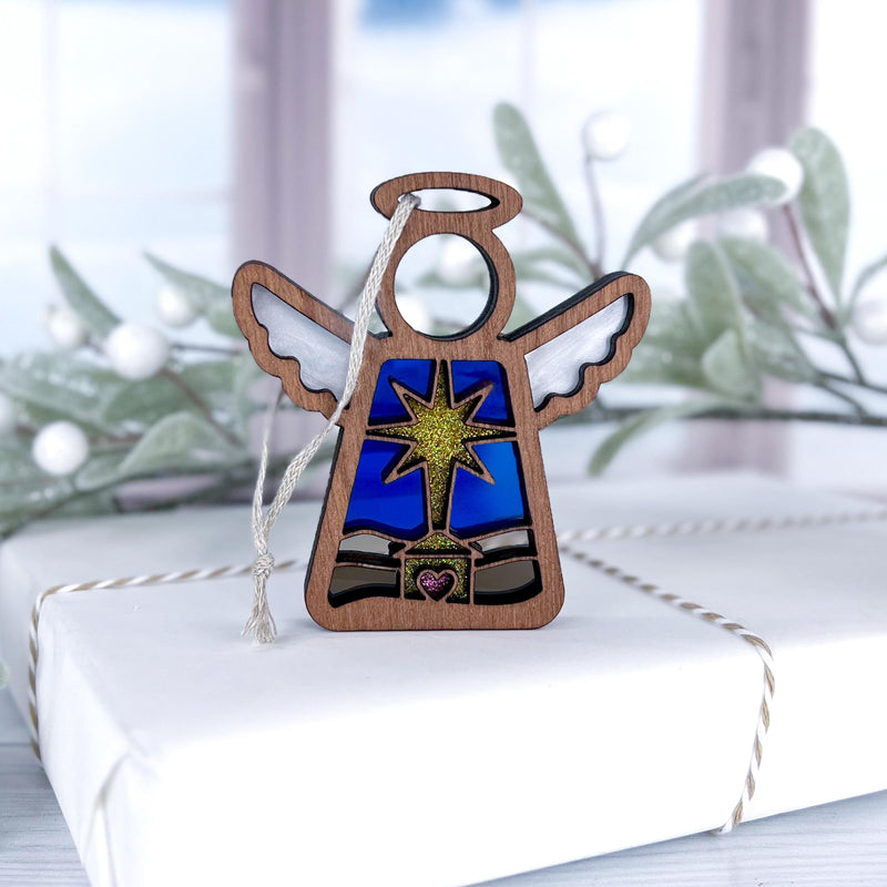 The Forged Flare® Christmas Nativity Star with Imperfections 3.5" Mother's Angels® wooden angel figurine sits on a twine-tied gift box, surrounded by softly blurred holiday décor and frosted greenery for a heartfelt holiday scene.