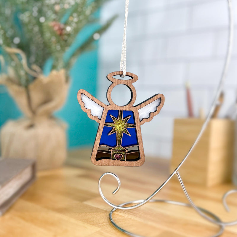 The Forged Flare® Christmas Nativity Star 3.5" (H) Angel Figurine from Mother's Angels® sits on a wooden table, hanging from a silver stand, with a burlap-wrapped potted plant and festive decor in the blurred background.