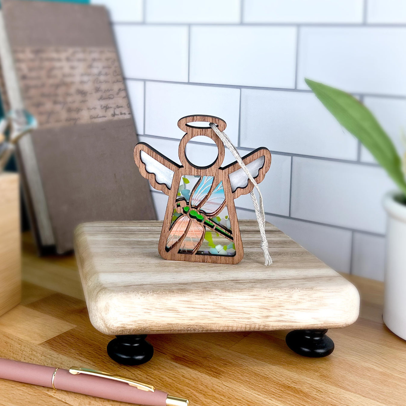 The 3.5" Mother's Angels® Dragonfly Ornament by Forged Flare® features wood and stained glass with a tassel on a small stand, ideal for gifting or finding as a Warehouse Return on Amazon. In the background are a notebook, plant, and pink pen on a wooden desk.