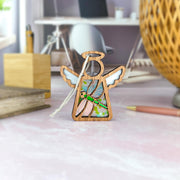 A Forged Flare® Warehouse Return - Dragonfly Ornament | 3.5" Mother's Angels® Angel Figurine with stained glass wings stands on a pink desk with stationery, books, and a lamp in a softly lit, modern workspace—ideal for Amazon shoppers.
