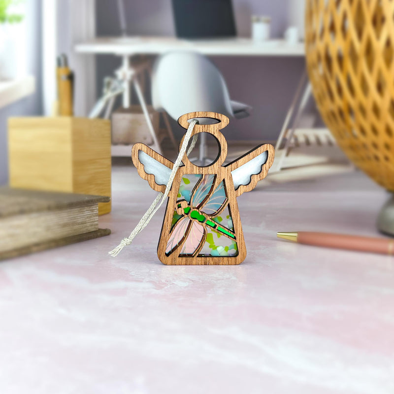 A Forged Flare® Warehouse Return - Dragonfly Ornament | 3.5" Mother's Angels® Angel Figurine with stained glass wings stands on a pink desk with stationery, books, and a lamp in a softly lit, modern workspace—ideal for Amazon shoppers.