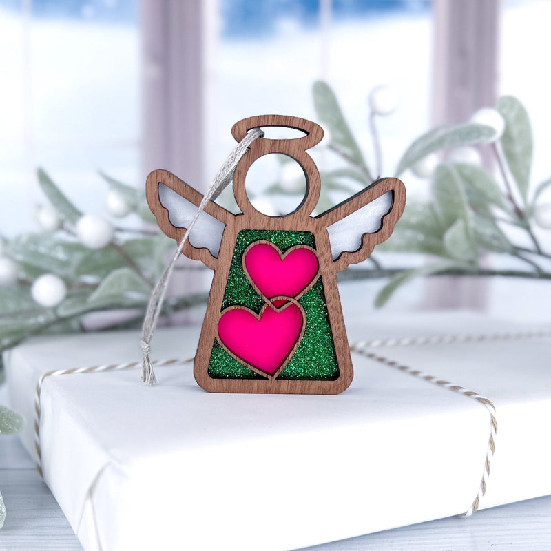 The Forged Flare® Limited Edition Interlocking Hearts Ornament – Series 8, a 3.5" Mother’s Angels® figurine, is displayed on a white gift box with frosted greenery and a snowy window in the background.