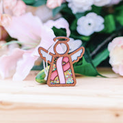 The Forged Flare® Mother's Angels® 3.5" Angel Figurine features a pink ribbon for breast cancer awareness and colorful beads, making it a thoughtful charity ornament gift. Blurred pink and white flowers appear in the background.