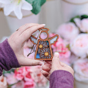 A person in a purple sweater holds a Forged Flare® Warehouse Return - Daisy Ornament, a 3.5" Mother's Angels® angel figurine, while pink and white flowers blur softly in the background, reminiscent of delightful Amazon finds.