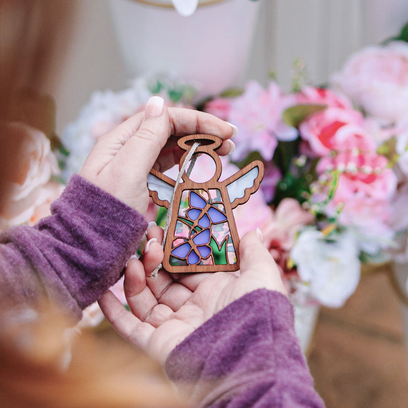 A person in a purple sweater holds a 3.5" Playful Purple Butterflies Angel Figurine from the Mother's Angels® collection by Forged Flare®, standing before pink and white flowers.