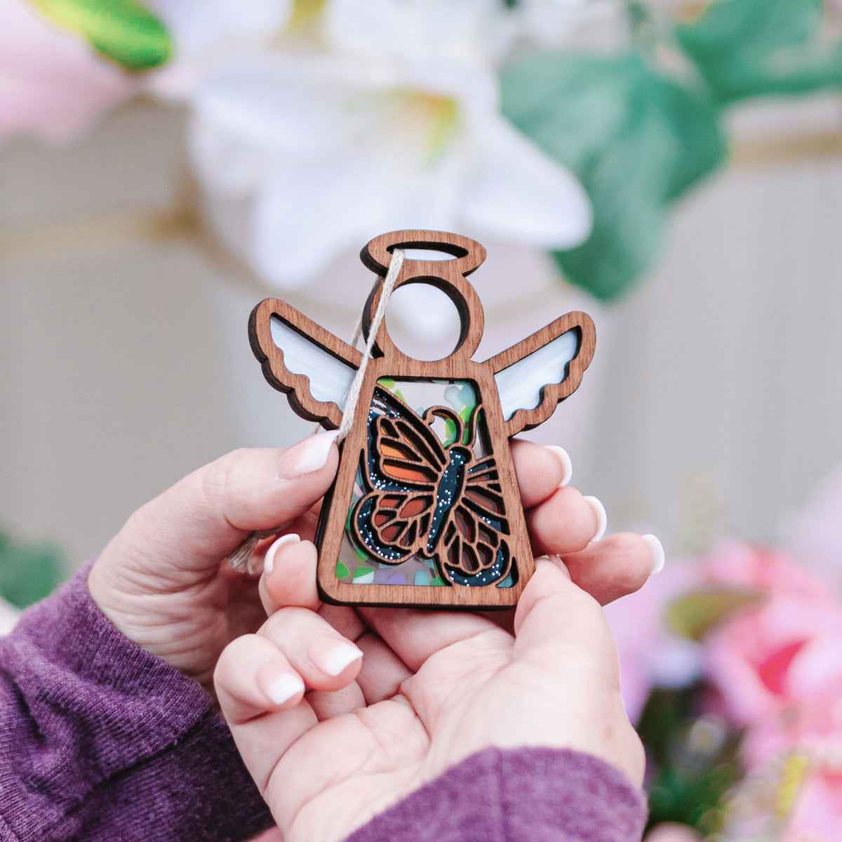 Close-up of hands holding a Forged Flare® Mother's Angels® Monarch Butterfly Ornament with Imperfections—a 3.5" angel figurine—against a floral background, making a touching memorial gift.