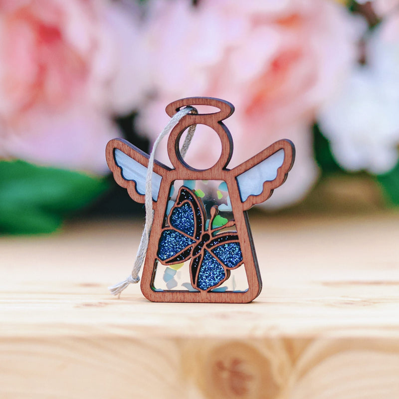 The Forged Flare® Mother's Angels® Morpho Blue Butterfly Ornament is a 3.5" wooden angel figurine with blue glitter wings, pictured on a light wood surface with a soft pink and green floral background. This item is a warehouse return.