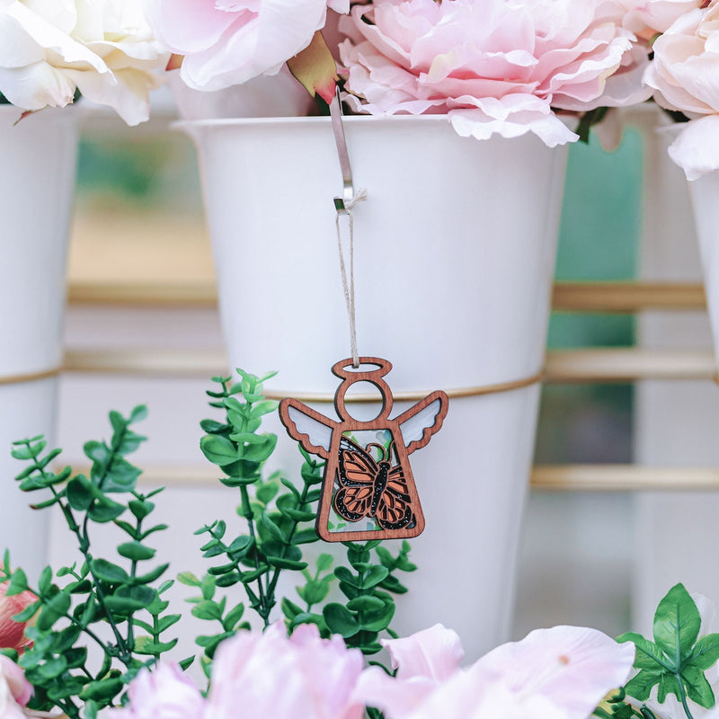 The Forged Flare® Mother's Angels® Monarch Butterfly Ornament with Imperfections, a 3.5" angel figurine, hangs among green leaves and pink flowers in white pots—an ideal memorial gift to cherish loved ones.
