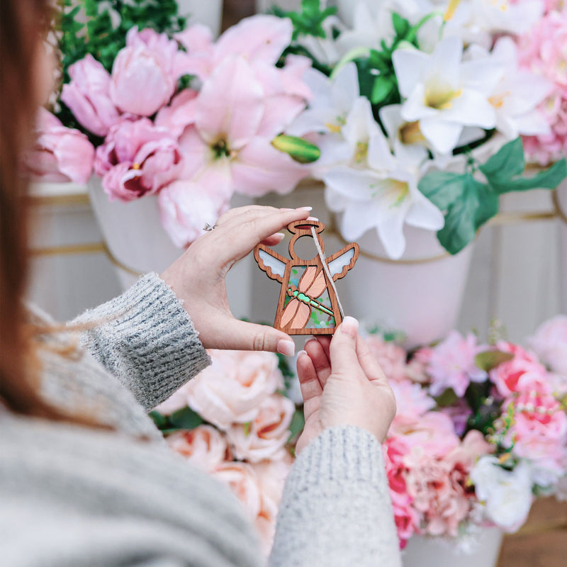 A person in a gray sweater holds a 3.5" Mother's Angels® Dragonfly Ornament by Forged Flare®, featuring white wings and flowers, standing before pink and white artificial flowers in pots. Ideal for gifting or shopping on Amazon as a warehouse return.