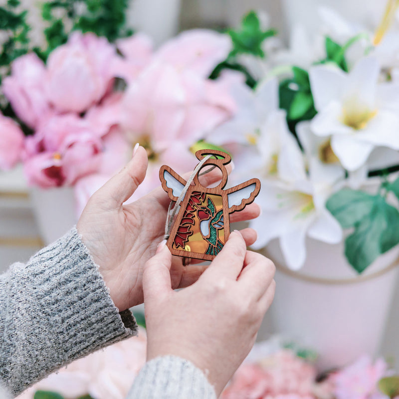 A person holds a Forged Flare® Mother’s Angels® Ruby-Throated Hummingbird Ornament (3.5" angel figurine), featuring colorful painted details, surrounded by white lilies and pink flowers, like those listed as warehouse returns on Amazon.