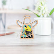 The Forged Flare® Goldfinch Ornament | 3.5" (H) Mother's Angels® figurine stands on a light wood surface—a charming bird lover gift. A blurred white pot with green leaves and flowers decorates the background.