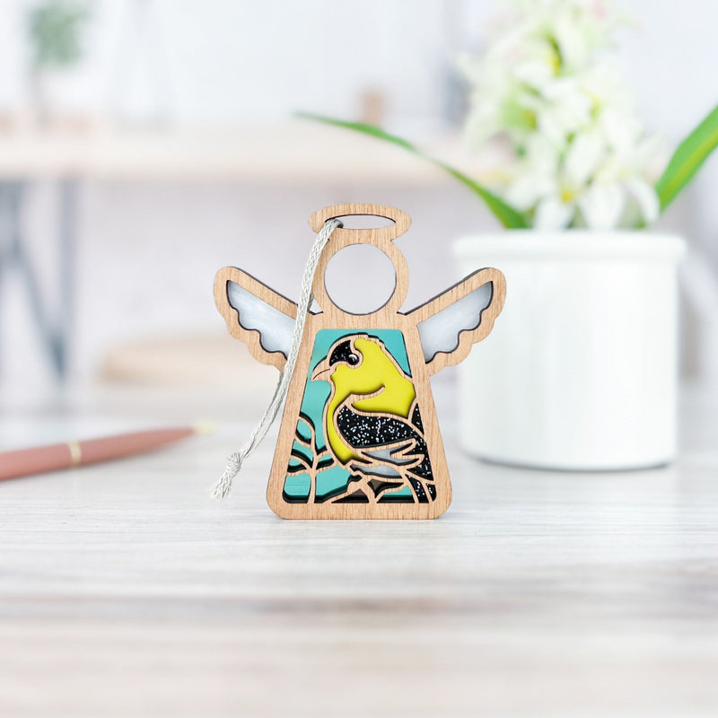 The Forged Flare® Goldfinch Ornament | 3.5" (H) Mother's Angels® figurine stands on a light wood surface—a charming bird lover gift. A blurred white pot with green leaves and flowers decorates the background.