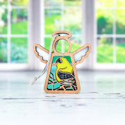 The Forged Flare® Goldfinch Ornament is a 3.5" Mother's Angels® angel figurine with a painted bird design, displayed on a light surface against blurred greenery—perfect as a gift for bird lovers.