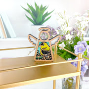 The Forged Flare® Goldfinch Ornament, a 3.5" Mother's Angels® angel figurine, stands on a gold shelf amid green and purple potted plants and white flowers, creating a bright, decorative scene for bird lovers.