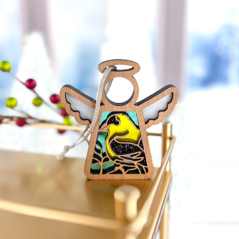 The Forged Flare® Goldfinch Ornament (3.5" H Mother's Angels® angel figurine) features stained glass-style wings and a goldfinch design, hanging by a string on a gold surface with festive decorations blurred in the background.