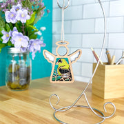 The Forged Flare® Goldfinch Ornament | 3.5" (H) Mother’s Angels® angel figurine hangs from a silver stand on a wood countertop, perfect for bird lovers. A jar of purple flowers and a wooden container with pencils sit in the background.