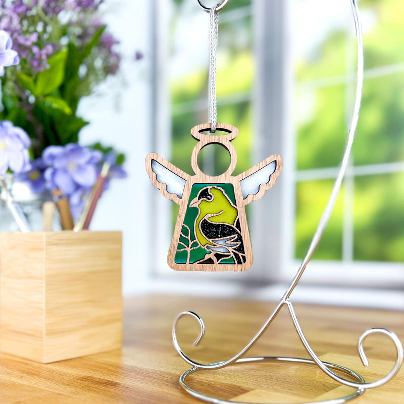 The Forged Flare® Goldfinch Ornament | 3.5" Angel Figurine from Mother's Angels® features a bird design and hangs from a metal stand—an ideal bird lover gift, shown with purple flowers and sunlight streaming through the window.