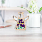 The Forged Flare® Iris Ornament, a 3.5" Mother's Angels® angel figurine with a stained glass-style purple iris design, stands on a light wood surface—ideal for flower décor or angel collections. A blurred flower pot and pen are in the background.