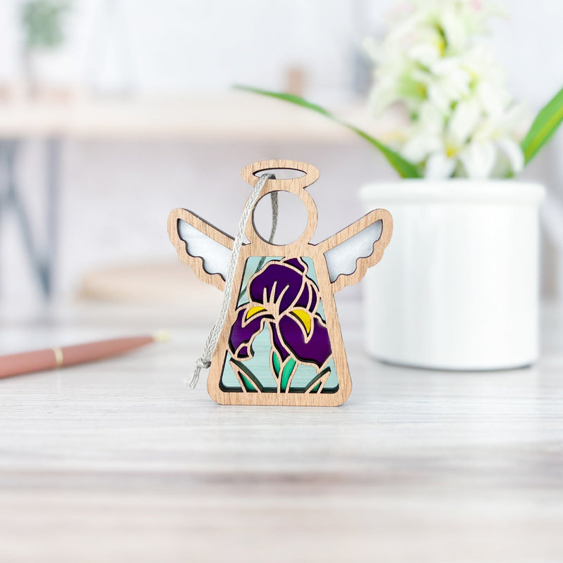 The Forged Flare® Iris Ornament, a 3.5" Mother's Angels® angel figurine with a stained glass-style purple iris design, stands on a light wood surface—ideal for flower décor or angel collections. A blurred flower pot and pen are in the background.