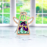 The Forged Flare® Iris Ornament is a 3.5" Mother's Angels® figurine with stained glass-style wings and a purple iris design, displayed on a white surface before sunlit windows with garden views—a charming addition to any collection.