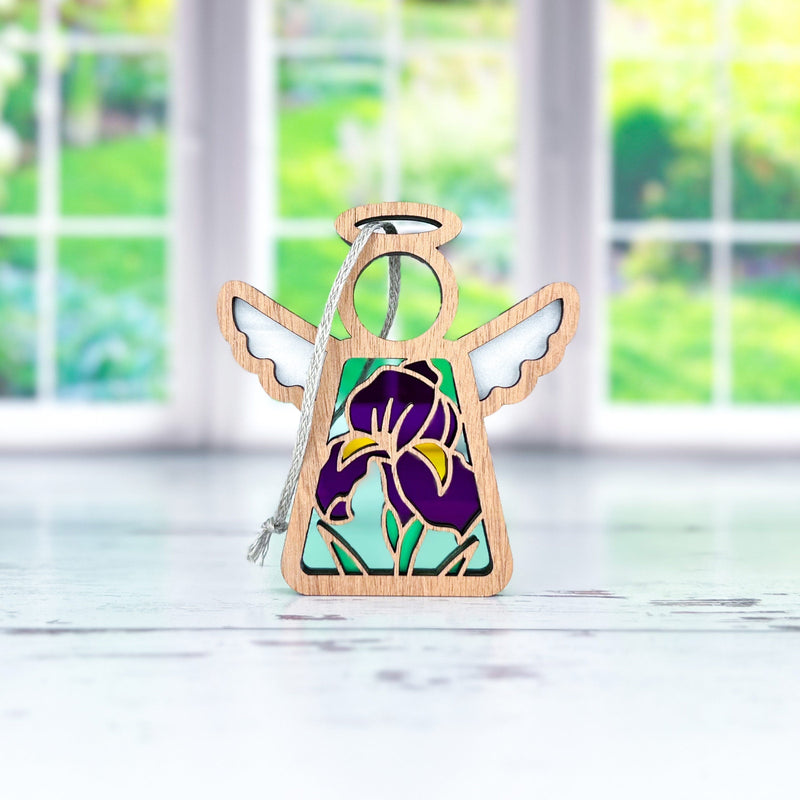 The Forged Flare® Iris Ornament is a 3.5" Mother's Angels® figurine with stained glass-style wings and a purple iris design, displayed on a white surface before sunlit windows with garden views—a charming addition to any collection.