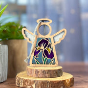 The Forged Flare® Iris Ornament, a 3.5" Mother's Angels® figurine with stained glass wings and purple flower design, stands on wood slices—a charming addition to any angel or floral decor. A potted plant and blue background complete the scene.