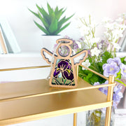 The Forged Flare® Mother's Angels® Iris Ornament, a 3.5" wooden angel figurine, stands on a gold shelf with green plants, books, and white and purple flowers in the background for a bright floral decor setting.