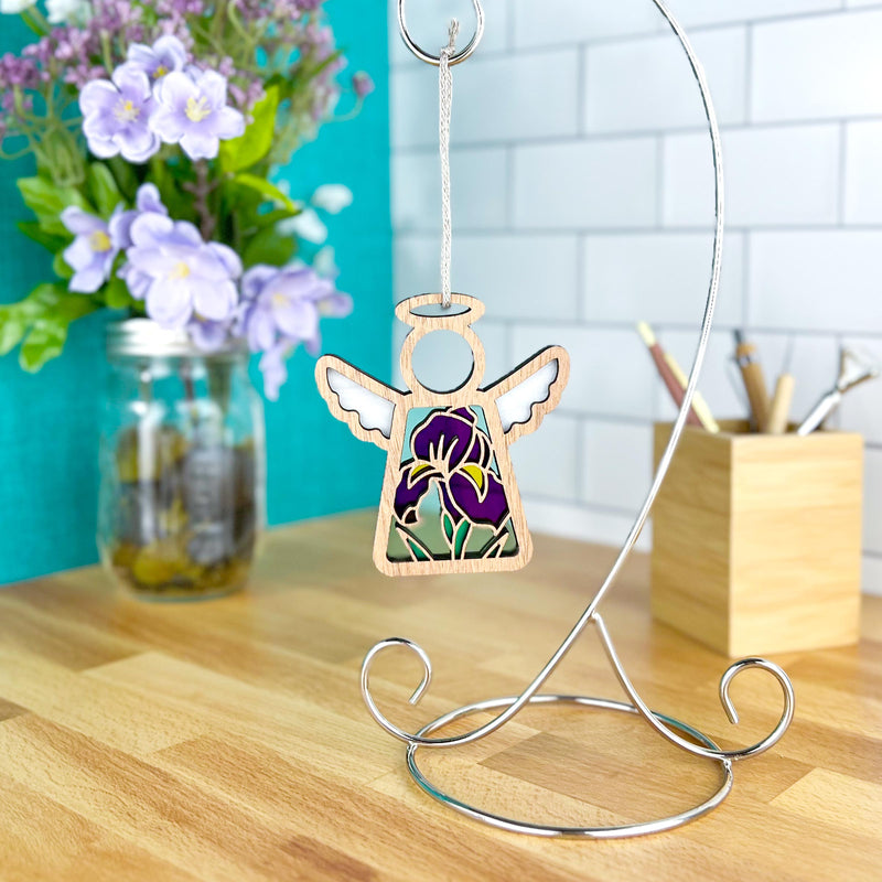 The Forged Flare® Iris Ornament | 3.5" (H) Angel Figurine from Mother's Angels® with purple and green glass hangs on a silver stand atop a wooden desk, with a jar of purple flowers and a wooden pencil holder in the background.
