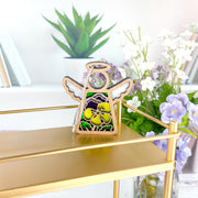 The Forged Flare® Pansy Ornament is a 3.5" Mother's Angels® figurine with stained glass-style wings and a yellow flower, displayed on a gold shelf with books, a potted plant, and purple and white flowers in the background.