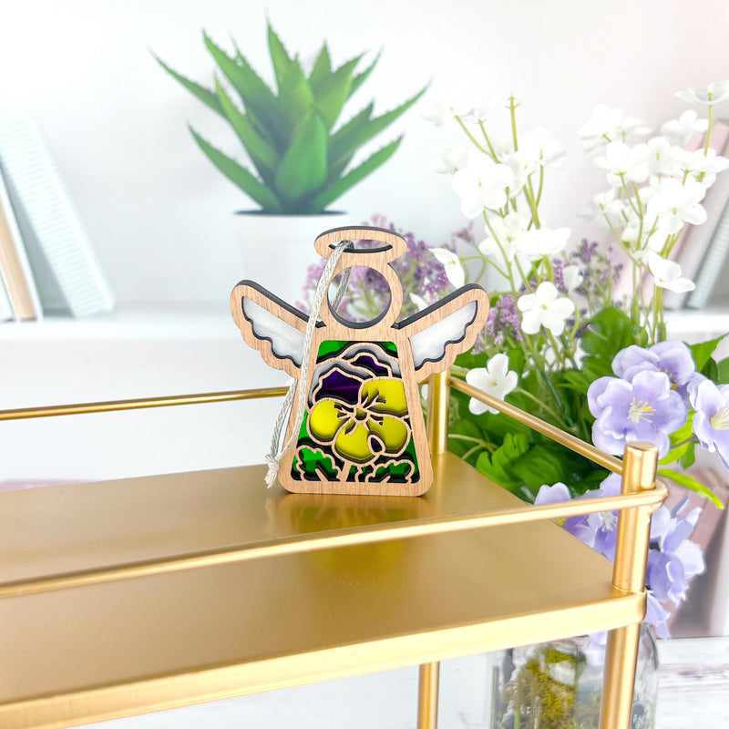 The Forged Flare® Pansy Ornament is a 3.5" Mother's Angels® figurine with stained glass-style wings and a yellow flower, displayed on a gold shelf with books, a potted plant, and purple and white flowers in the background.