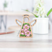 The Forged Flare® Mother's Angels® Pink Evening Primrose Ornament is a 3.5" wooden angel figurine with green leaves, pink flowers at the center, and a silver string, standing on a light wood surface with a blurred mug and plant behind it.