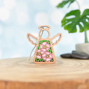 The Forged Flare® Pink Evening Primrose Ornament | 3.5" Angel Figurine from Mother's Angels® sits on a light wood surface, with soft-focus stones and a potted plant in the background. The angel features wings, a halo, pink flowers, and green leaves.