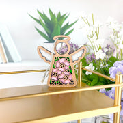 The Forged Flare® Pink Evening Primrose Ornament, a 3.5" Mother's Angels® figurine, stands on a gold shelf with books and green and white potted plants adding charm in the background.