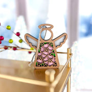 The Forged Flare® Pink Evening Primrose Ornament is a 3.5" Mother's Angels® figurine featuring pink primrose flowers and green leaves, standing on a gold surface with a silver hanging string and festive lights in the blurred background.