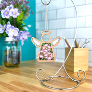The Forged Flare® Pink Evening Primrose Ornament, a 3.5" Mother's Angels® angel figurine, hangs on a silver stand on the kitchen counter, with a jar of purple flowers and pencils in a wooden holder in the background.