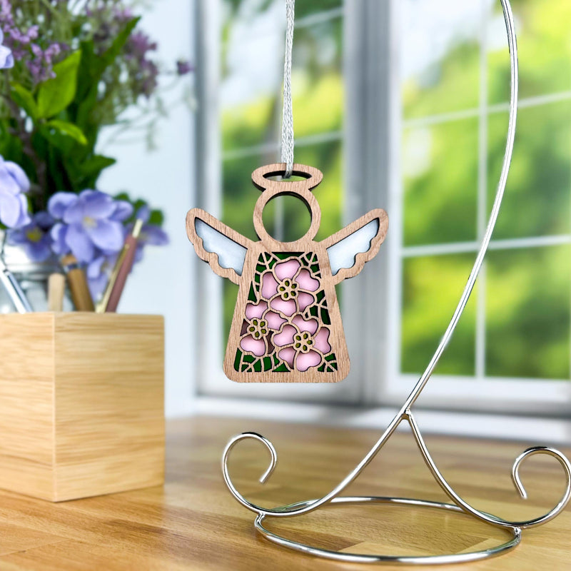 The Forged Flare® Mother's Angels® Pink Evening Primrose Ornament, a 3.5" wooden angel figurine, hangs from a decorative metal stand on a wooden desk with purple flowers and a window in the background.