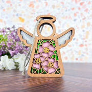 The Forged Flare® Pink Evening Primrose Ornament, a 3.5" wooden angel figurine from Mother's Angels®, features floral cutouts, pink accents, and a twine loop for hanging on wood with blurred purple and white flowers in the background.