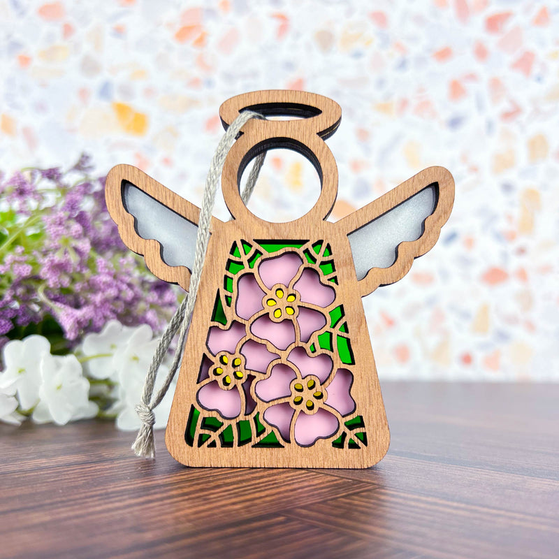 The Forged Flare® Pink Evening Primrose Ornament, a 3.5" wooden angel figurine from Mother's Angels®, features floral cutouts, pink accents, and a twine loop for hanging on wood with blurred purple and white flowers in the background.