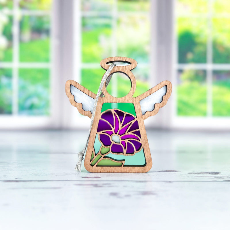 The Forged Flare® Mother's Angels® Purple Morning Glory Ornament, a 3.5" wooden angel with white wings and a floral birth flower design, stands on a white surface with a silver string, in front of a bright window blurred by greenery.