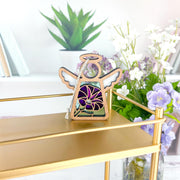 The Forged Flare® Purple Morning Glory Ornament | 3.5" (H) Angel Figurine from Mother's Angels® sits on a gold shelf, with books, a potted plant, and white and purple flowers providing a bright, modern backdrop.