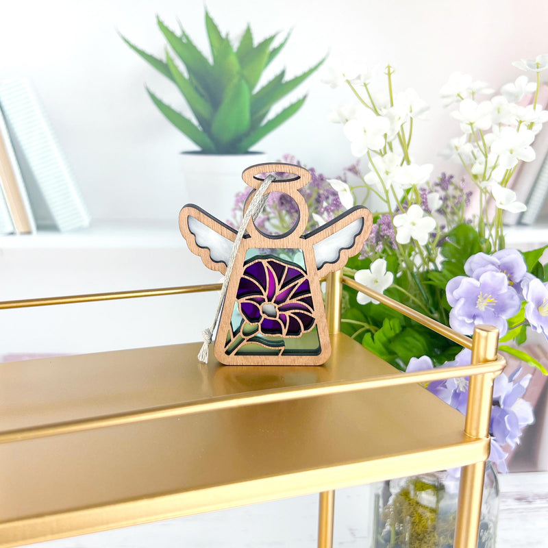 The Forged Flare® Purple Morning Glory Ornament | 3.5" (H) Angel Figurine from Mother's Angels® sits on a gold shelf, with books, a potted plant, and white and purple flowers providing a bright, modern backdrop.