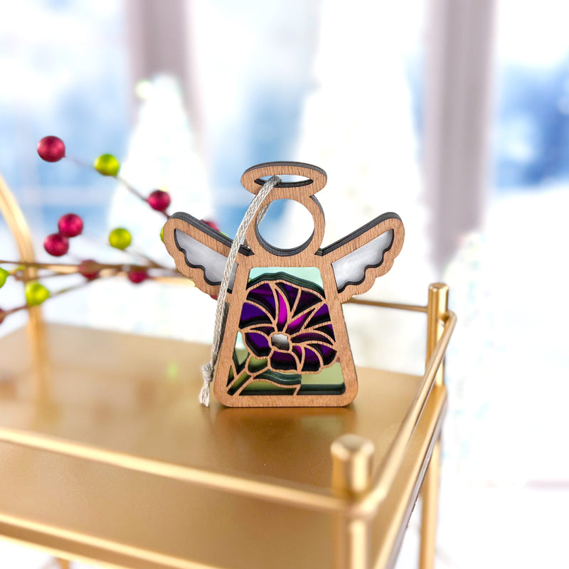 The Forged Flare® Purple Morning Glory Ornament, a 3.5" Mother's Angels® figurine with stained glass-style wings and a June birth flower design, hangs by a silver string on a golden shelf amid festive décor and a wintry blur.