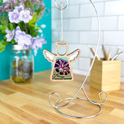 The Forged Flare® Purple Morning Glory Ornament, a 3.5" Mother's Angels® figurine, hangs on a metal stand atop a wooden countertop, with a jar of purple morning glories and a container of pencils in the background.