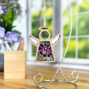A Forged Flare® Purple Morning Glory Ornament, a 3.5" Mother's Angels® angel figurine with white wings and a purple morning glory design, hangs from a metal stand on a table with blurred flowers and a window in the background.