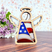 The Forged Flare® Mother's Angels® 3.5" American Flag Ornament features a wood angel figurine with stained glass stars and stripes, resting on wood with purple flowers—an eye-catching patriotic gift.