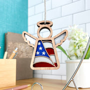 The Forged Flare® Mother's Angels® American Flag Ornament is a 3.5" wooden angel figurine with wings and a halo, hanging by a string amid office supplies, a white tile wall, and a potted plant in the background.