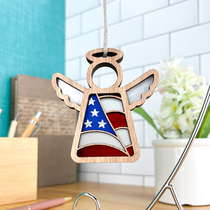 The Forged Flare® Mother's Angels® American Flag Ornament is a 3.5" wooden angel figurine with wings and a halo, hanging by a string amid office supplies, a white tile wall, and a potted plant in the background.