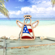 The Forged Flare® American Flag Ornament—a 3.5" Mother’s Angels® figurine—stands on a weathered table with seashells, set against a tropical beach backdrop, making a perfect patriotic gift.