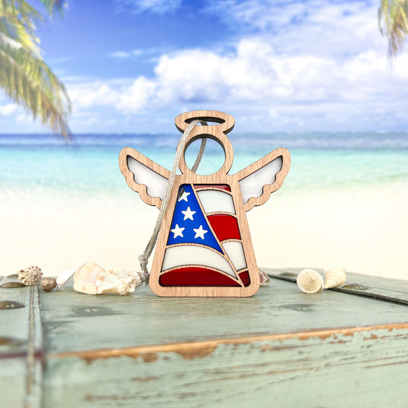 The Forged Flare® American Flag Ornament—a 3.5" Mother’s Angels® figurine—stands on a weathered table with seashells, set against a tropical beach backdrop, making a perfect patriotic gift.