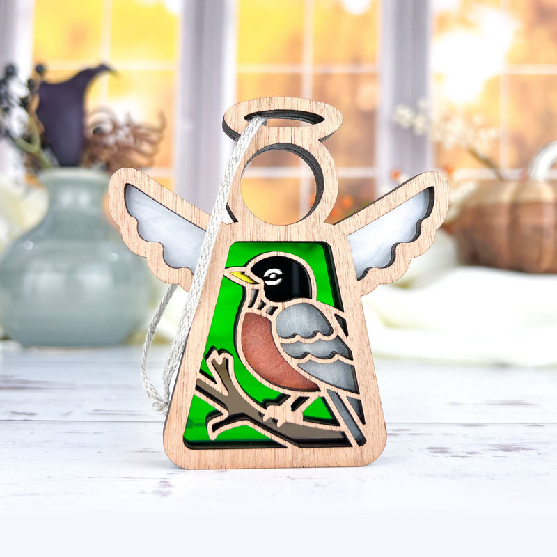 The Forged Flare® American Robin Ornament | 3.5" Mother's Angels® features a wooden angel with white wings and green stained glass, displaying a robin on a branch—perfect as a bird lover gift. Blurred vases and a window appear in the background.